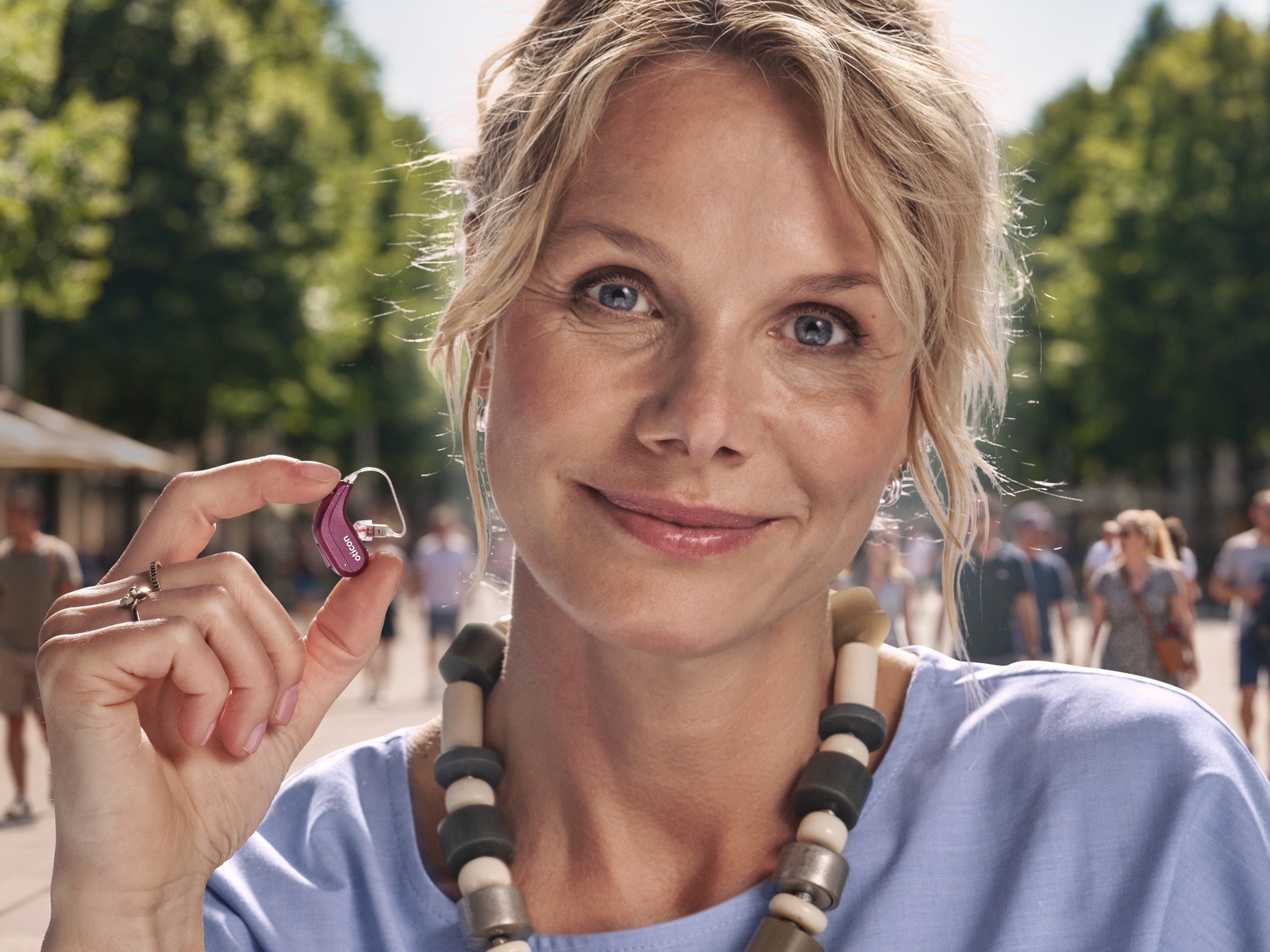 A woman on a sunny street holds the compact pink Oticon Reveal miniRITE hearing aid between her fingers