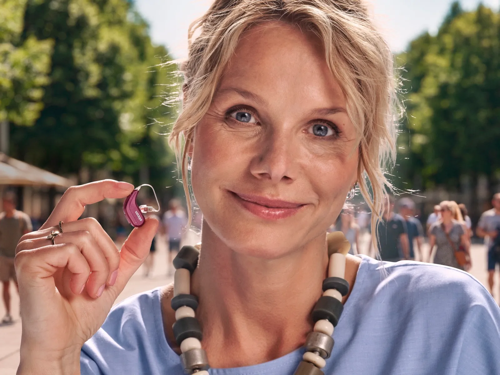 A woman on a sunny street holds the compact pink Oticon Reveal miniRITE hearing aid between her fingers