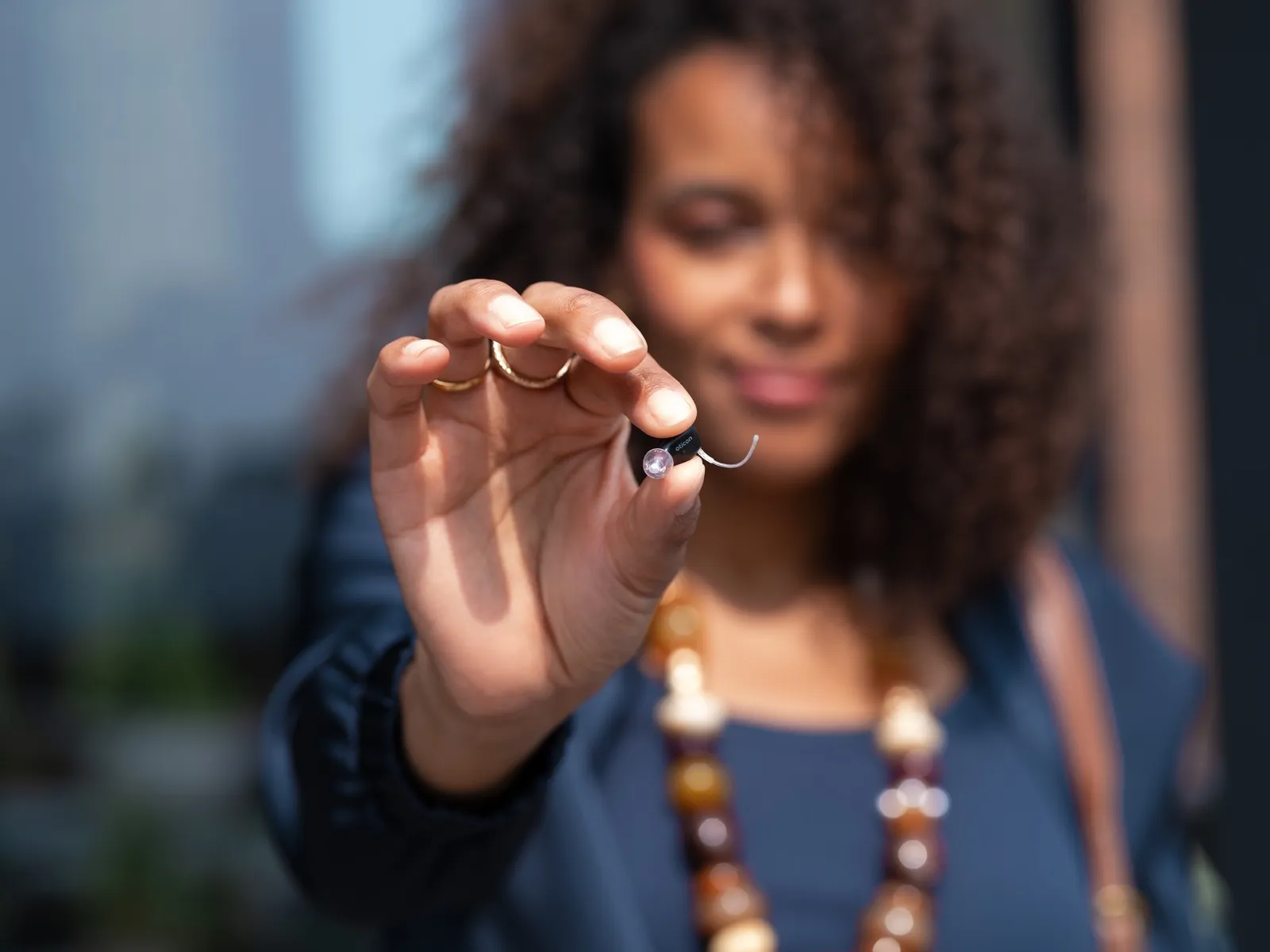 A woman holds the tiny Oticon Zeal in-the-ear hearing aid between two fingers, showing how small and discreet it is
